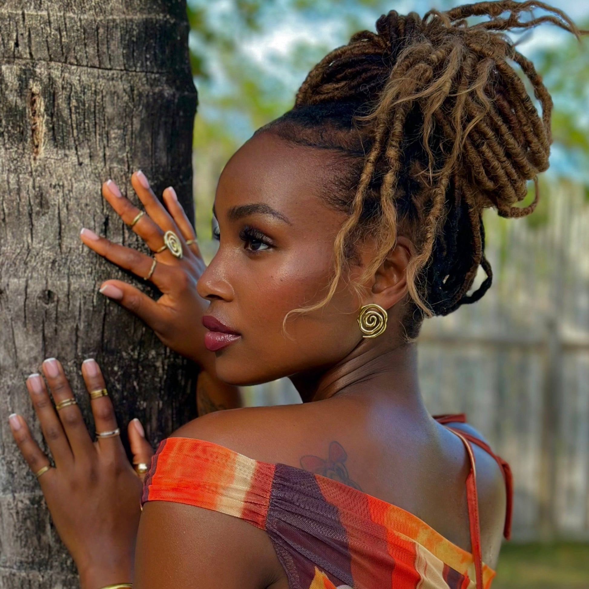 Woman with Loc braids leaning against a palm tree with gold spiral earrings.